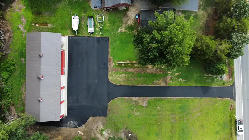 Aerial view of a freshly paved asphalt driveway widening into a parking apron beside a metal storage building