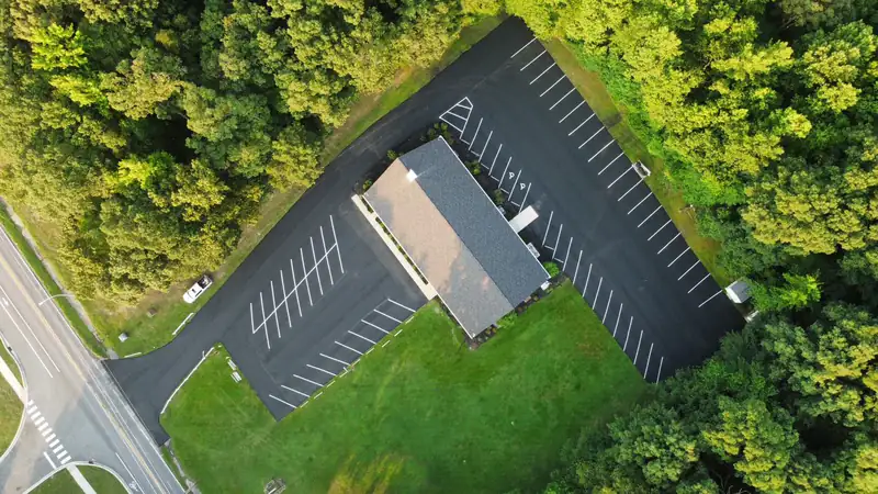 Aerial view of a freshly paved asphalt parking lot with new white striping surrounding a building beside hardwood trees