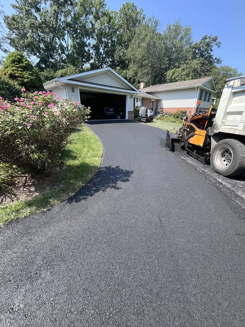 Asphalt paver and dump truck finishing a new blacktop driveway leading to a two-car garage at a split-level home