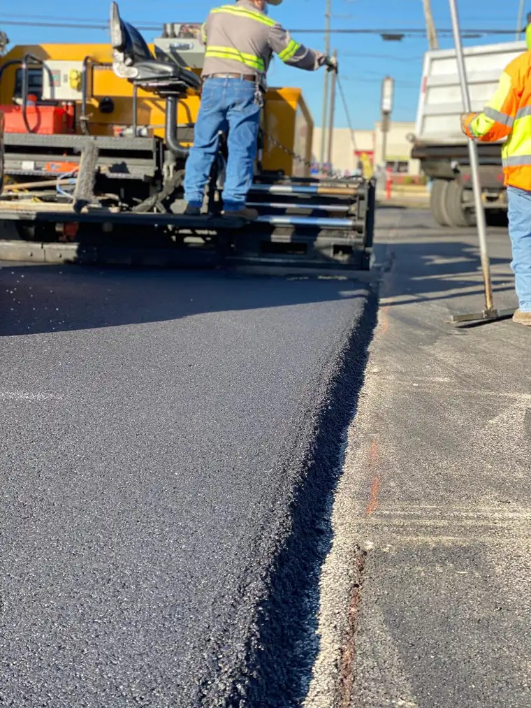 Asphalt paver laying a fresh hot-mix mat on a commercial lot as crew members work the edge