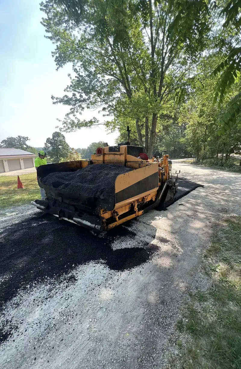 Asphalt paver loaded with hot mix paving over a white gravel country driveway under shade trees