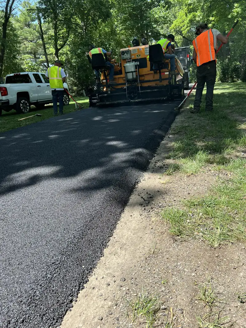 Asphalt paving crew laying and raking a fresh hot-mix driveway under mature shade trees