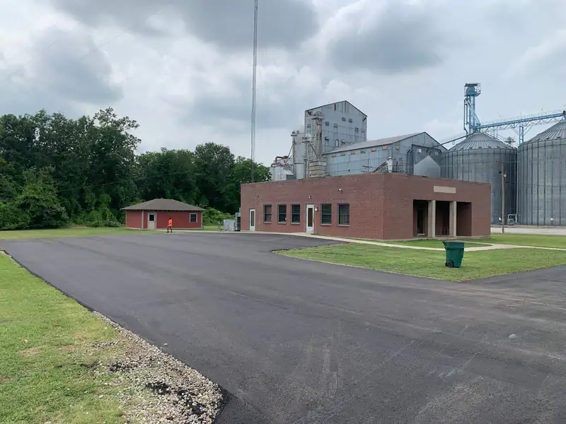Finished asphalt parking lot wrapping around a brick office building beside large steel grain bins