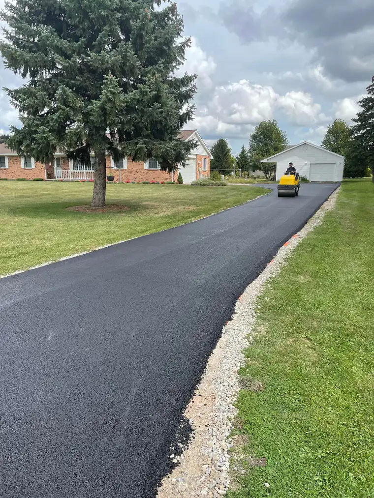 Freshly paved asphalt driveway with gravel shoulder leading to a brick ranch home and detached garage