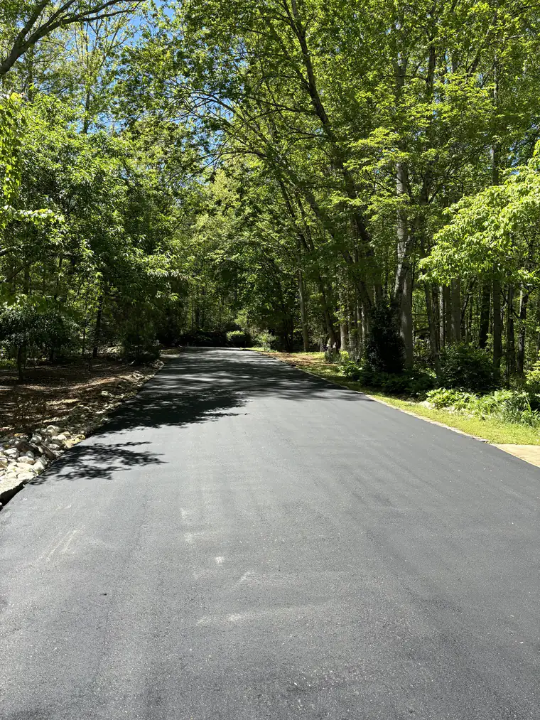 Freshly paved asphalt driveway winding through a shaded hardwood forest with river rock edging along one side
