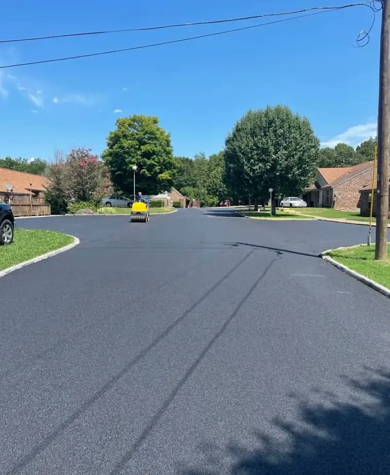 Freshly paved asphalt street through a brick townhome community, with a small roller compacting in the distance
