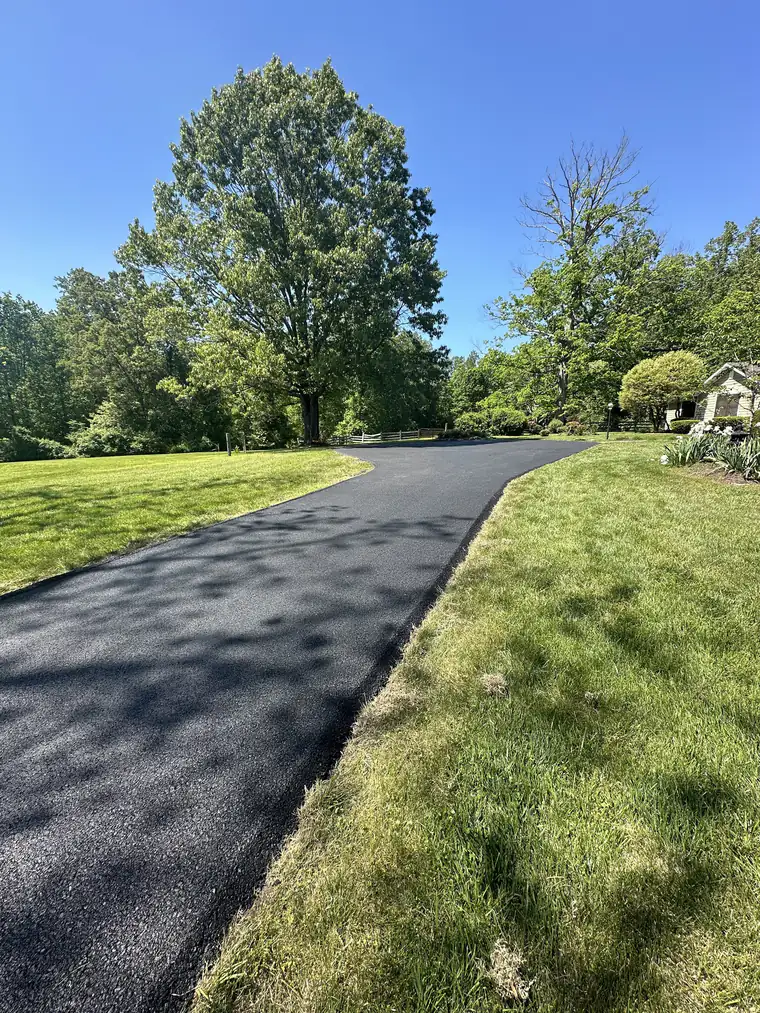 Freshly paved black asphalt driveway curving through a green lawn toward a large shade tree and split-rail fence