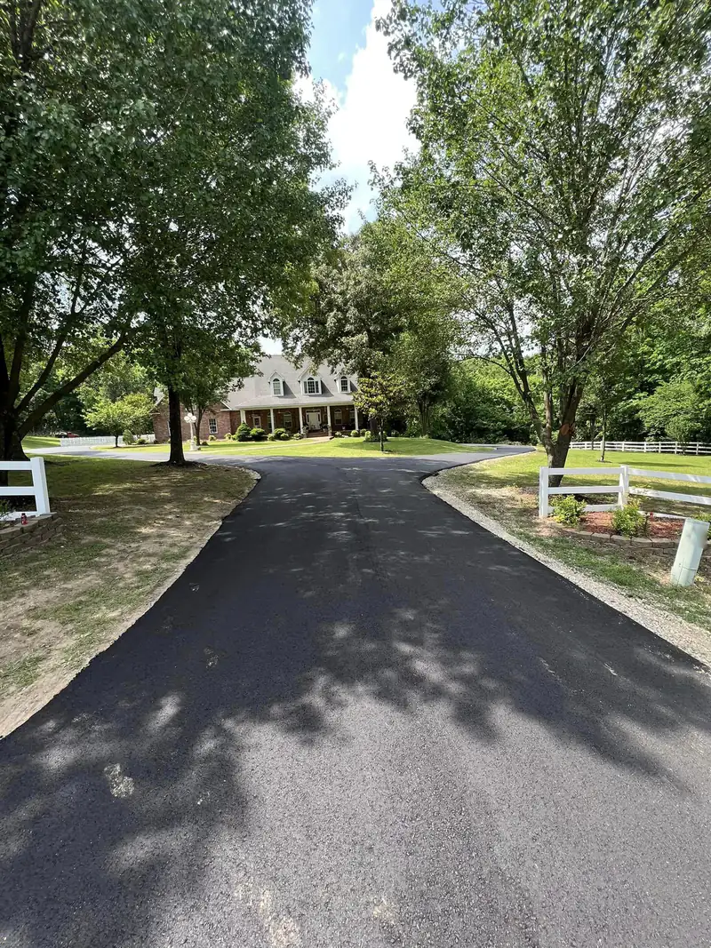 Freshly paved black asphalt driveway with gravel shoulders leading to a brick home under mature shade trees