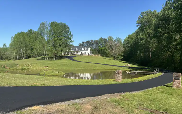 Freshly paved black asphalt driveway sweeping past a farm pond and brick entry pillars toward a white farmhouse on a wooded hill