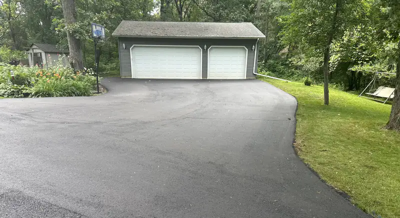 Freshly paved black asphalt driveway widening to a three-car detached garage, lined by green lawn and mature hardwood trees