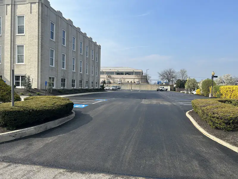 Freshly paved and striped asphalt parking lot beside a brick institutional building, taped off while it cures