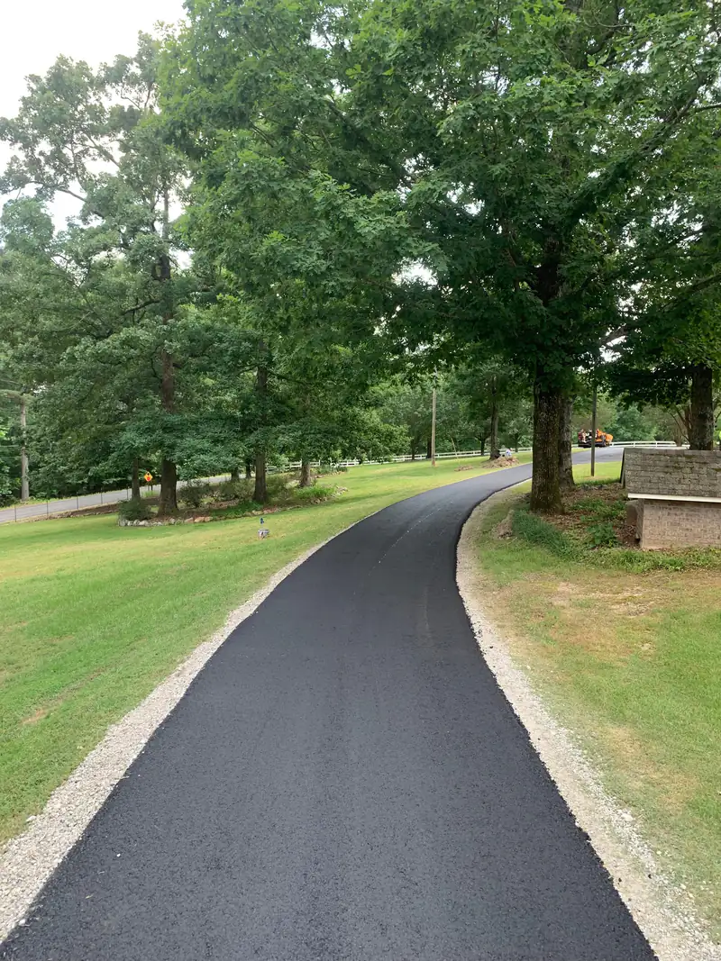 Freshly paved winding asphalt driveway with crushed stone edges curving beneath mature oak trees