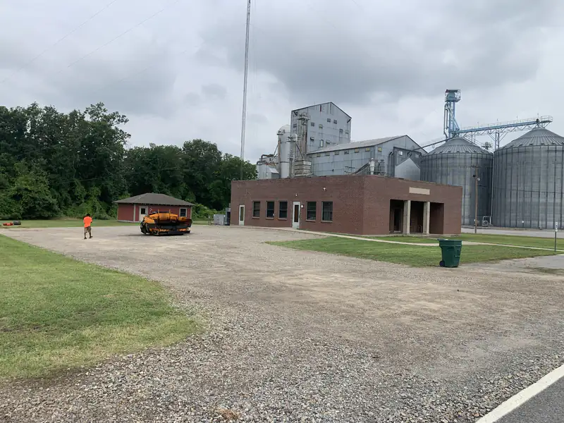 Gravel commercial lot before paving, with an asphalt paver staged beside a brick building and grain bins