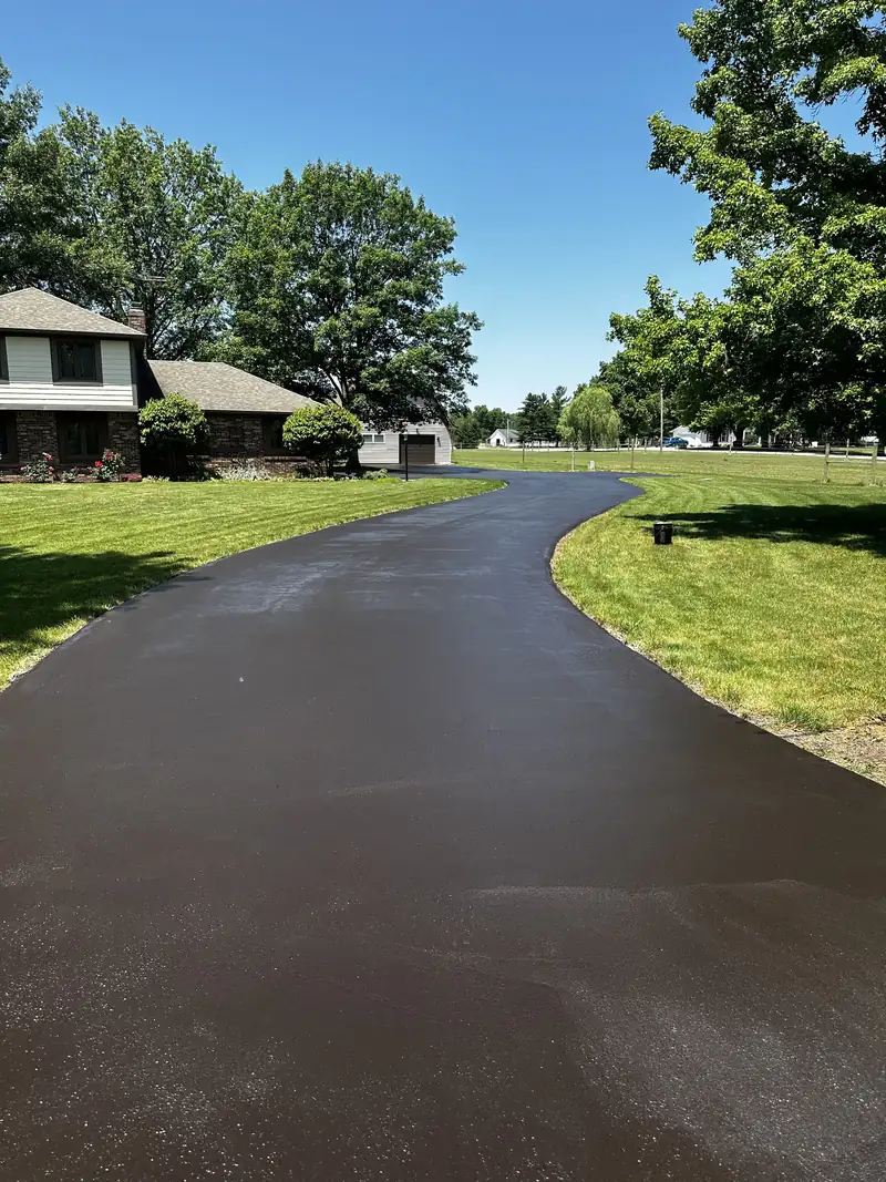 Long curving blacktop driveway, freshly sealcoated, passing a brick two-story home with mature shade trees