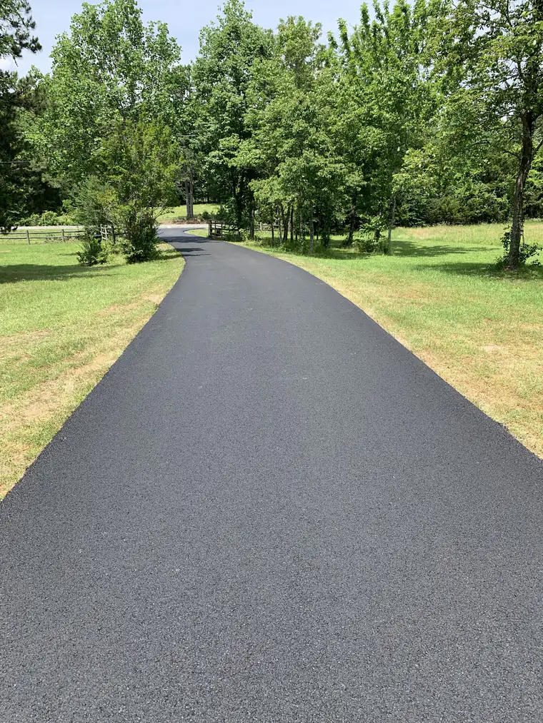 Long freshly paved black asphalt driveway curving through a mowed lawn toward a split-rail fence and shade trees