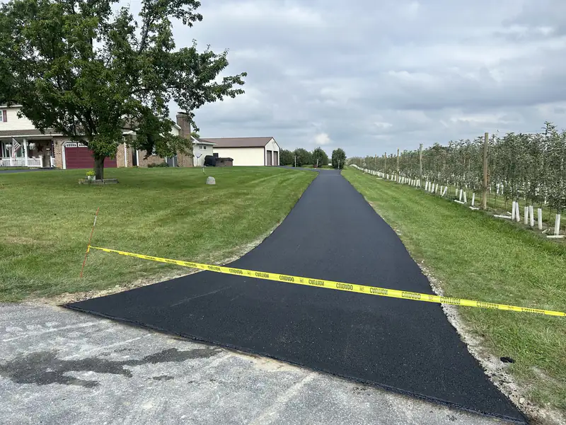 Long new asphalt driveway running past a brick ranch home and orchard toward a metal pole barn