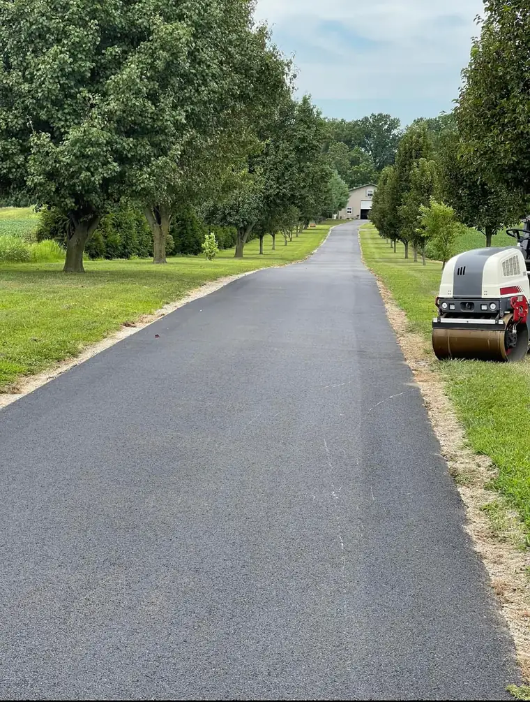 Long rural asphalt driveway lined with shade trees beside farm fields, with a small roller parked