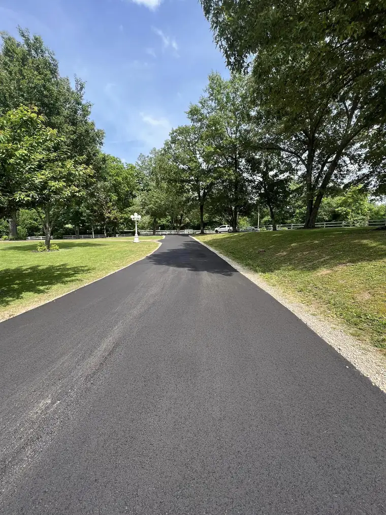Long tree-lined asphalt driveway with a fresh black surface, crushed stone edges and white rail fencing