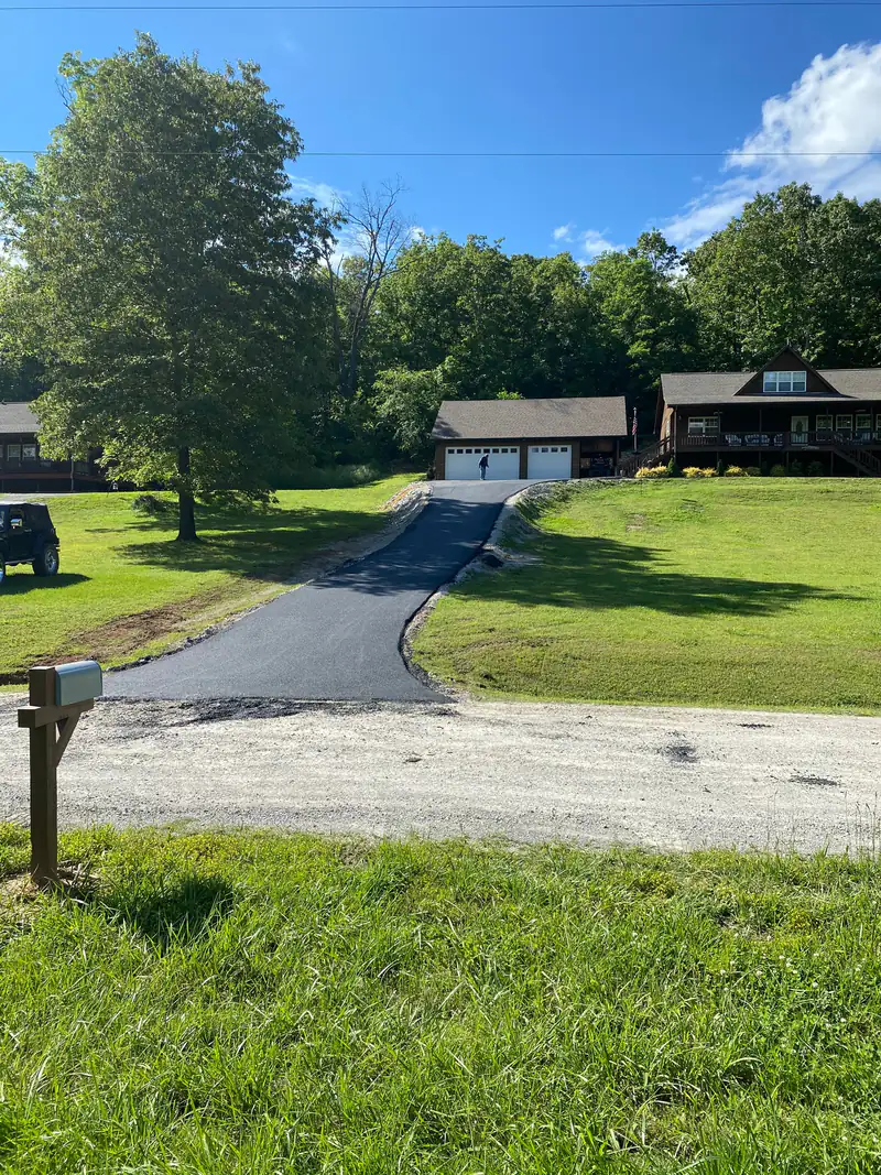 New asphalt driveway climbing a grassy hillside from a gravel road to a detached two-car garage