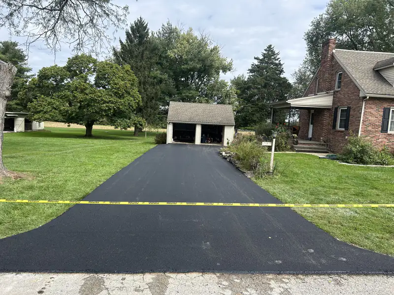 New asphalt driveway leading to a detached two-car garage beside a brick farmhouse and cornfield