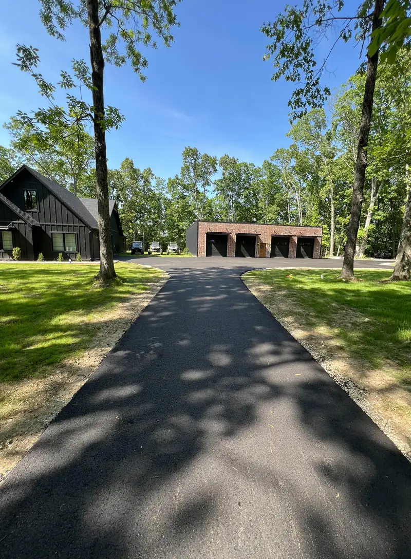 New asphalt driveway winding through tall oak trees to a brick four-bay garage and modern farmhouse
