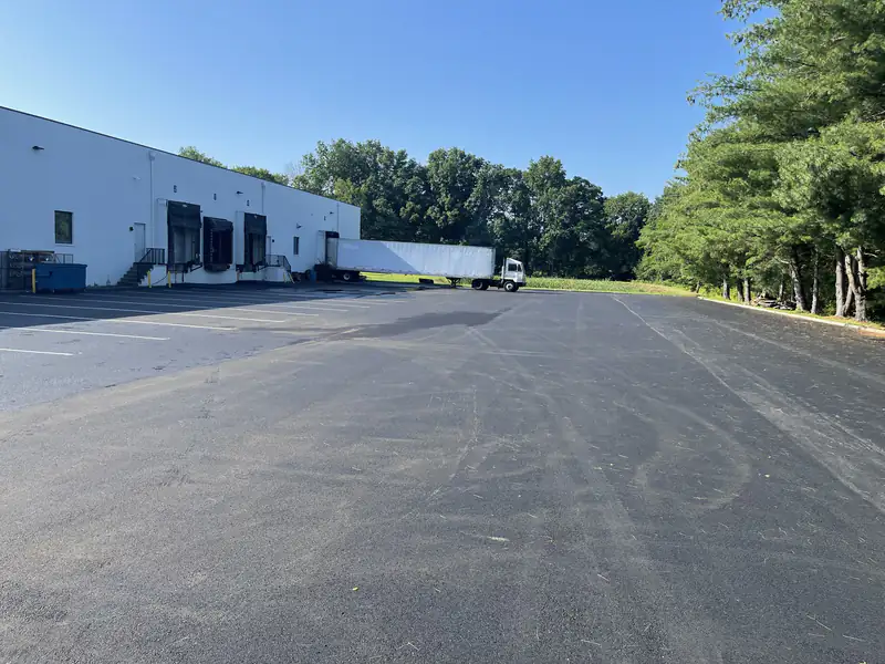 Newly paved asphalt lot behind a white warehouse with loading docks and a parked semi trailer.