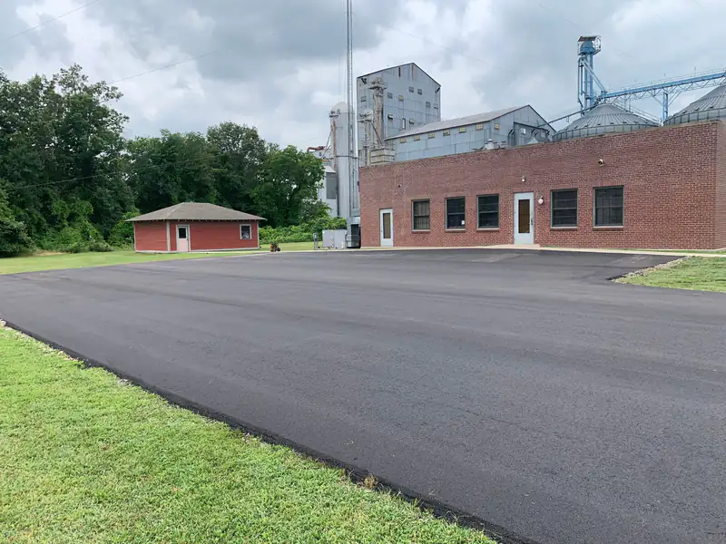 Newly paved asphalt parking lot beside a brick industrial building with a grain elevator and bins behind