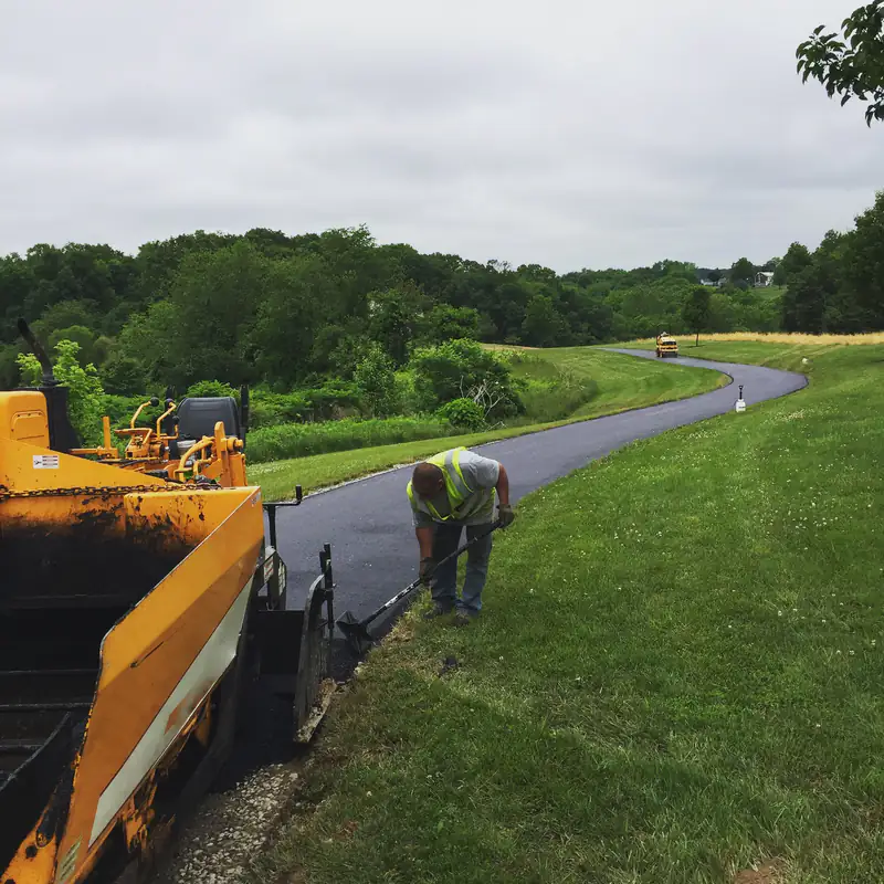 Paving crew member squaring the edge of a winding new asphalt driveway through rolling green farmland, with the paver in the foreground