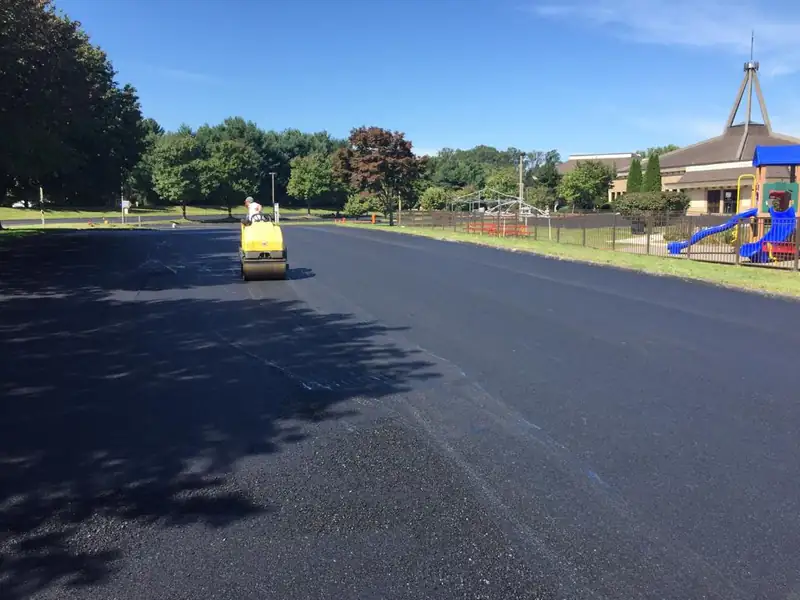 Ride-on roller compacting a freshly paved asphalt parking lot beside a church and fenced playground