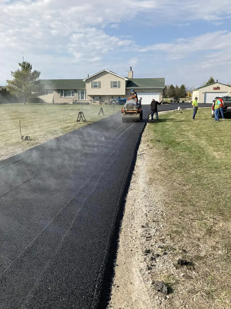 Ride-on roller compacting steaming fresh asphalt on a long rural driveway toward a split-level home