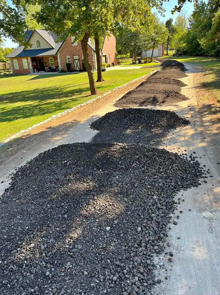 Rows of recycled asphalt millings delivered along a driveway beside a brick two-story home and oak trees