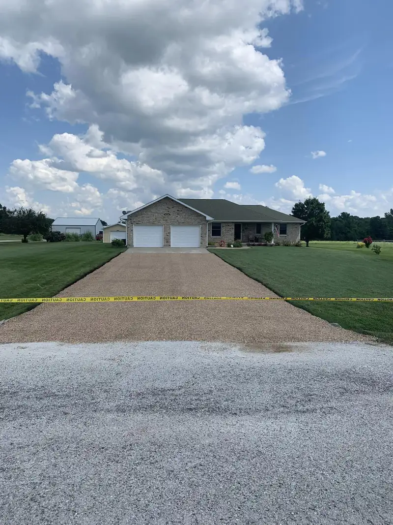 Tan chip seal driveway leading to a brick ranch home with a two-car garage and metal outbuilding