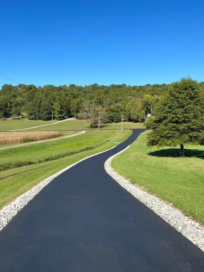 Winding black asphalt driveway with crushed limestone edging through green pasture, a cornfield and wooded hills