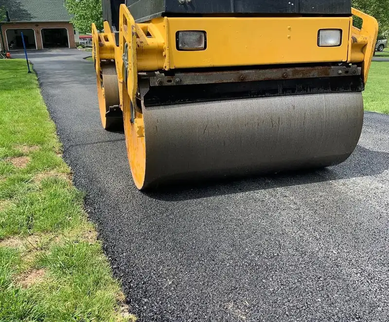 Yellow tandem roller drum compacting fresh asphalt on a residential driveway beside a green lawn